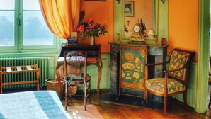 A room with a green wall, a wooden chair, a table with a vase of flowers, and a window with curtains.