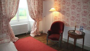 A room with a red chair, a small table with a lamp, a radiator, and floral wallpaper.