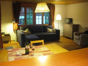 A furnished living room with a sofa, a dining table with a bottle and glasses, a television on a dresser, and a window with curtains.