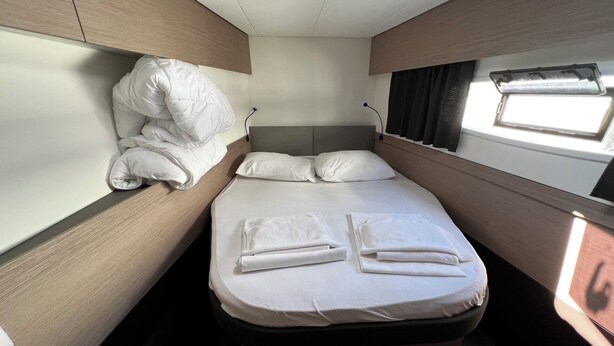 Cabine compacta e bem iluminada com uma cama de solteiro, roupa de cama e toalhas brancas impecáveis, cabeceira e janela com persianas.