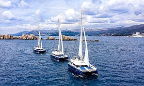 Three sailboats on calm waters with rocky outcrops and mountains in the background.