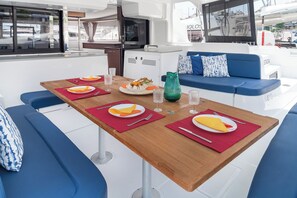 A dining area with a wooden table set for four, blue cushioned seating, and a view of the marina through large windows. - Sun-Kissed Sailing Adventure in Islas Sarónicas (Piraeus)