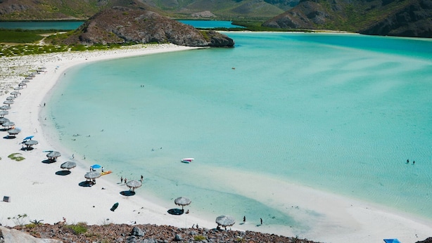 Uma praia com águas turquesa cristalinas, areia branca e colinas rochosas ao fundo.