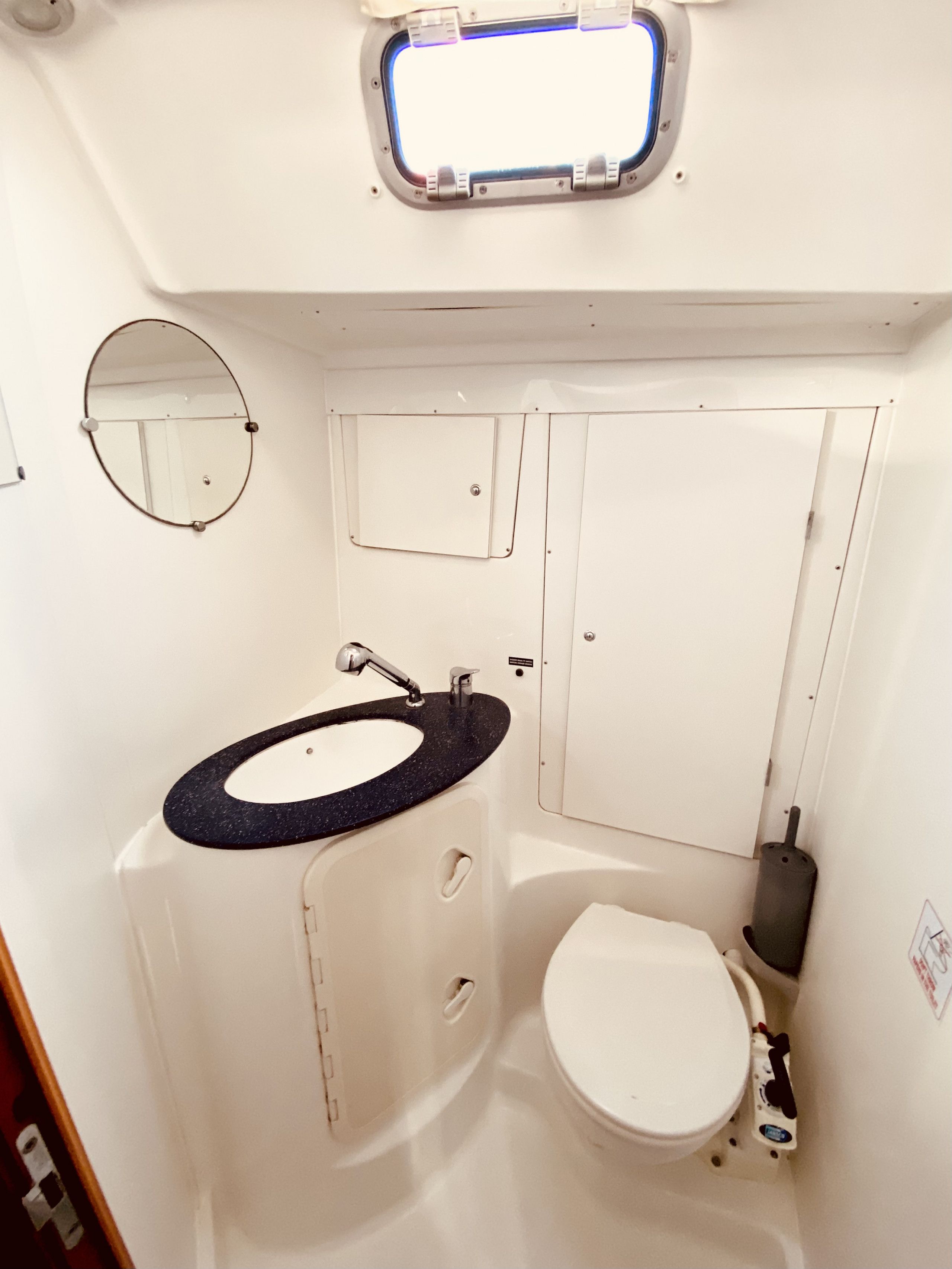 A compact bathroom with a round mirror, a sink with a black countertop, a toilet, and a small window above the sink.