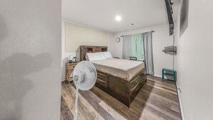 Un dormitorio con cama, mesita de noche, ventilador, reloj y ventana con cortinas.