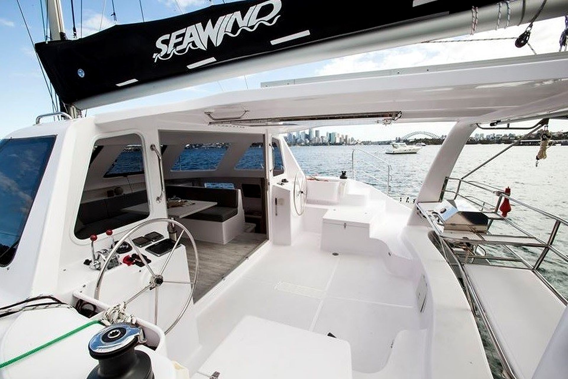 A sailboat's deck with a steering wheel, a seating area, and a view of the water and city skyline.