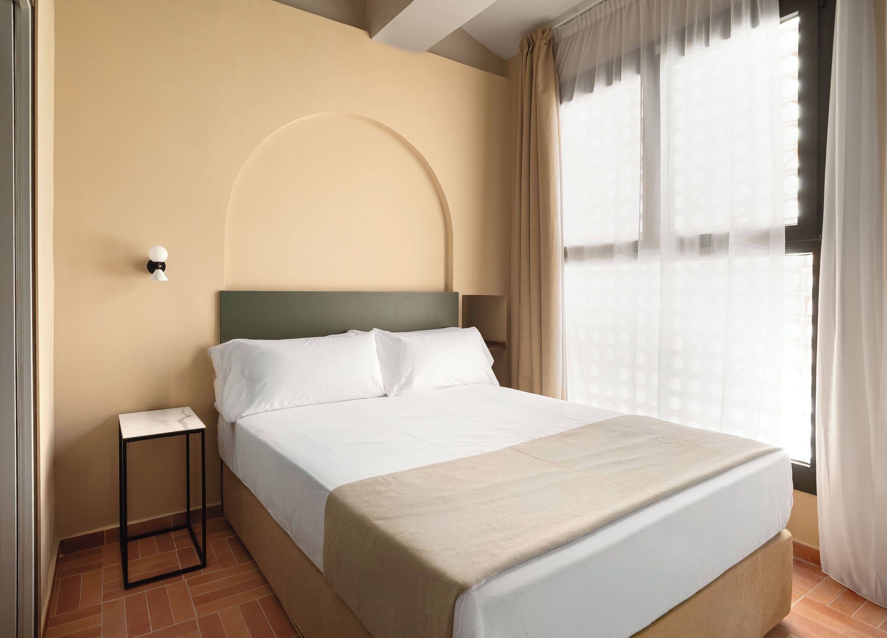 Habitación de hotel con cama, mesita de noche, lámpara de pared y ventana con cortinas.