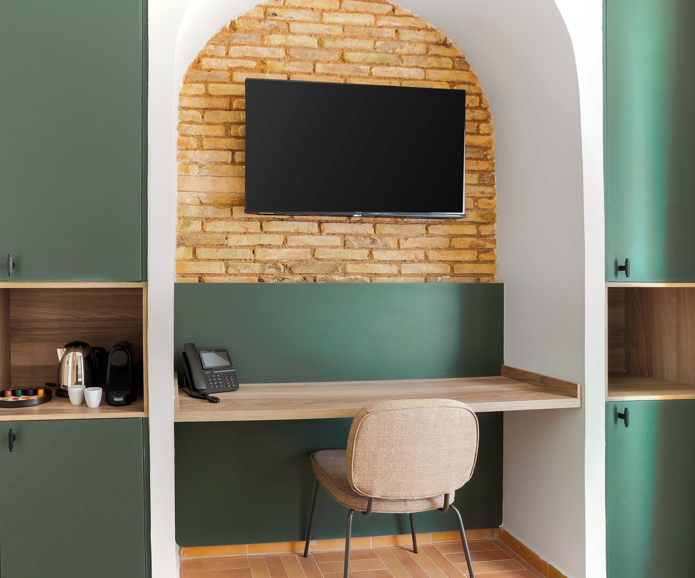 Een kamer met een bakstenen muur, een wandgemonteerde flatscreen-tv, een houten bureau, een rieten stoel en een koffiezetapparaat.