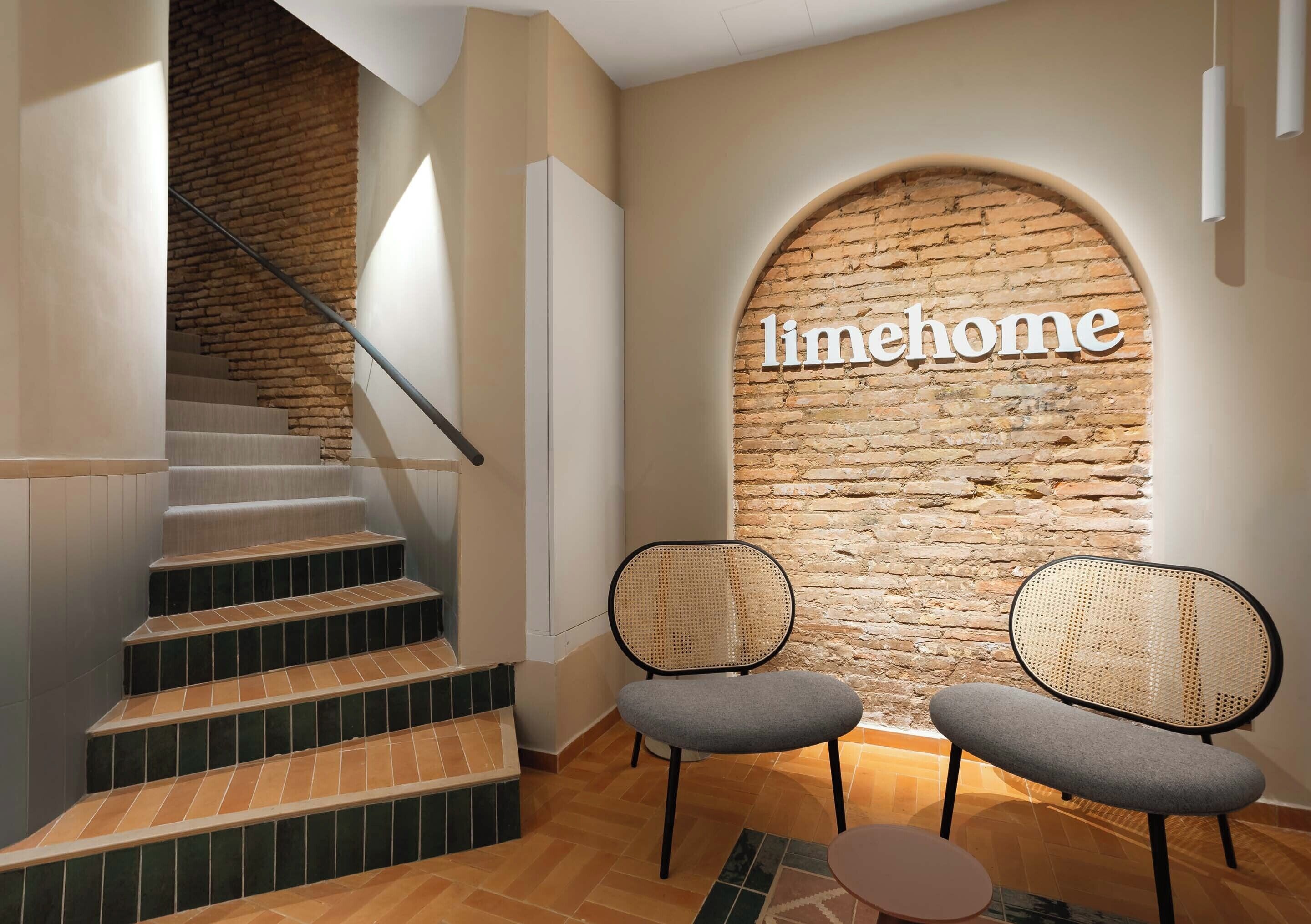 A staircase leading to a room with a brick wall and the word 'limehome' on it. There are two chairs and a small table in the room.