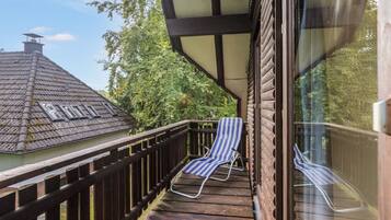Eine Holzterrasse mit zwei gestreiften Liegestühlen, einer Glastür und Blick auf ein Haus mit Ziegeldach.