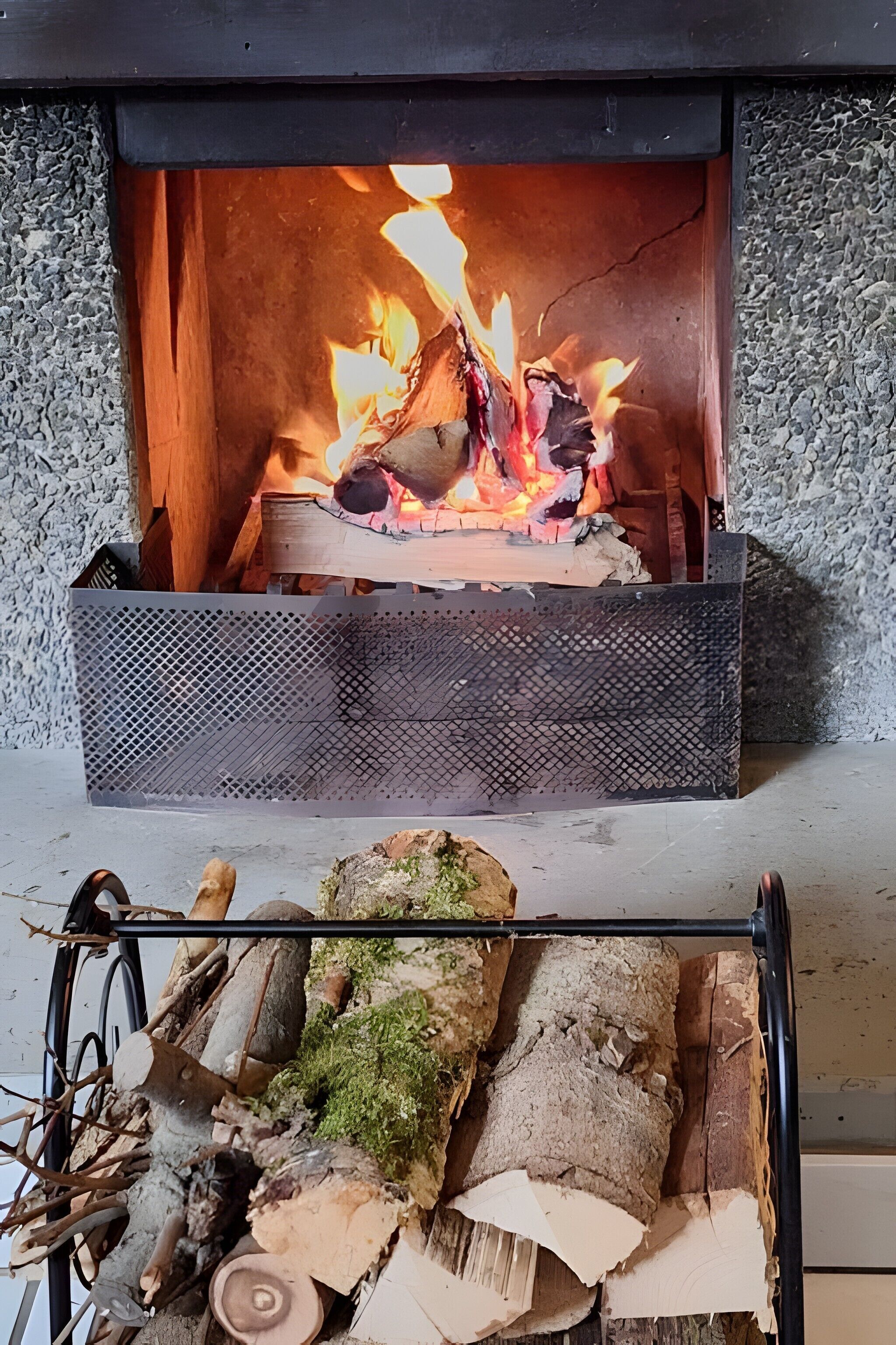 A fireplace with burning logs, a metal grate, and a stack of firewood outside.