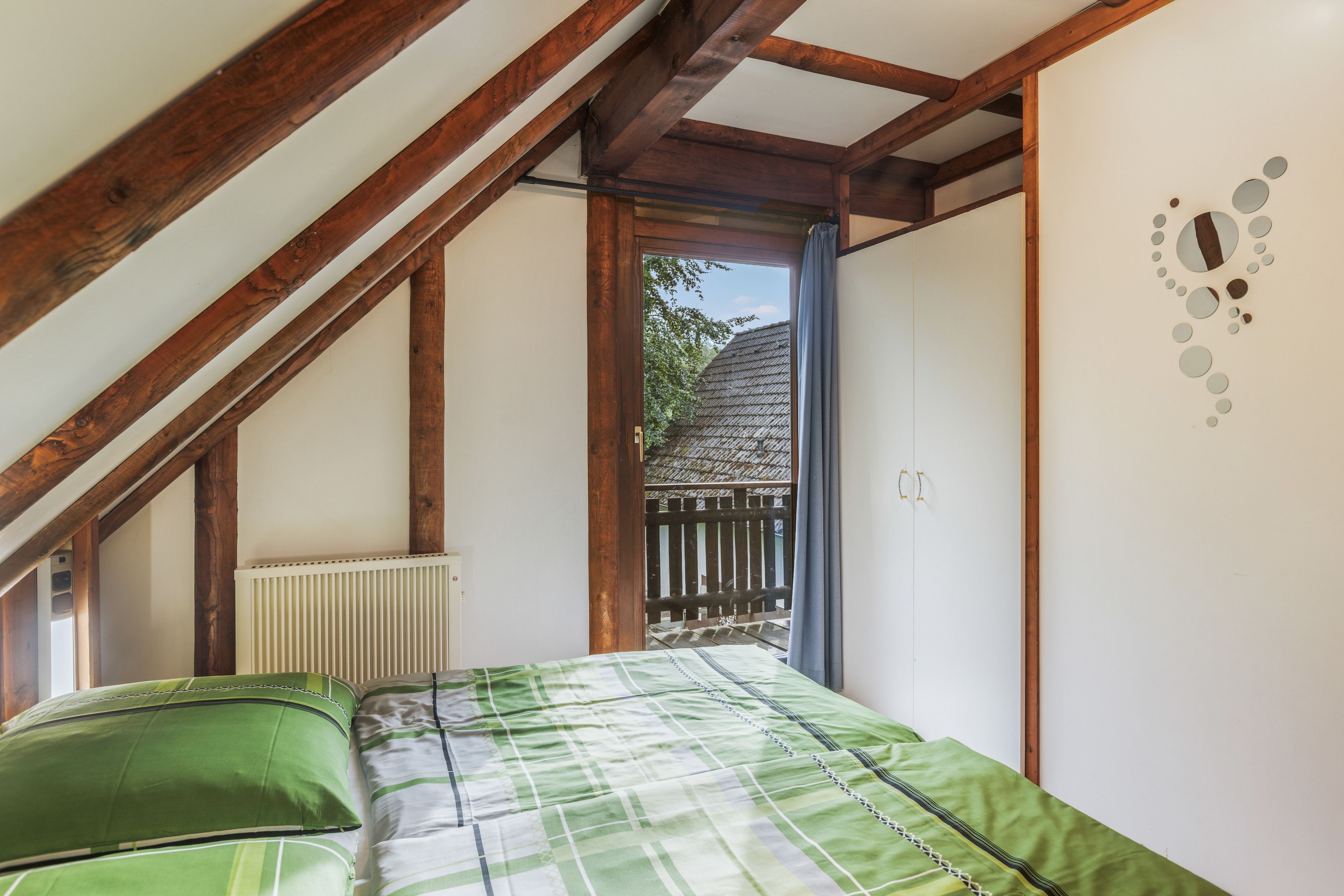 Une chambre avec un plafond en pente, un radiateur, un lit avec une literie verte et une vue sur une clôture en bois et des arbres à l’extérieur.