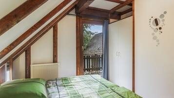Une chambre avec un plafond en pente, un radiateur, un lit avec une literie verte et une vue sur une clôture en bois et des arbres à l’extérieur.