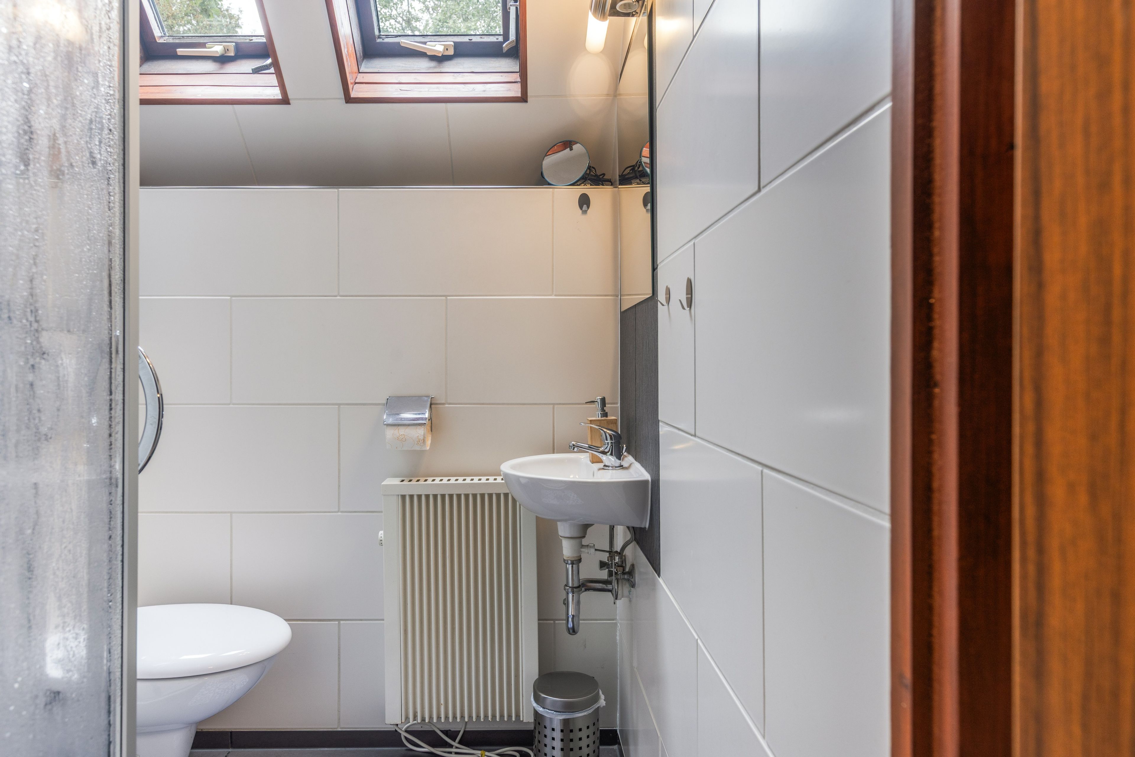 Ein kleines Badezimmer mit Toilette, Waschbecken, Heizkörper und Mülleimer. Oberhalb der Toilette befinden sich Dachfenster.