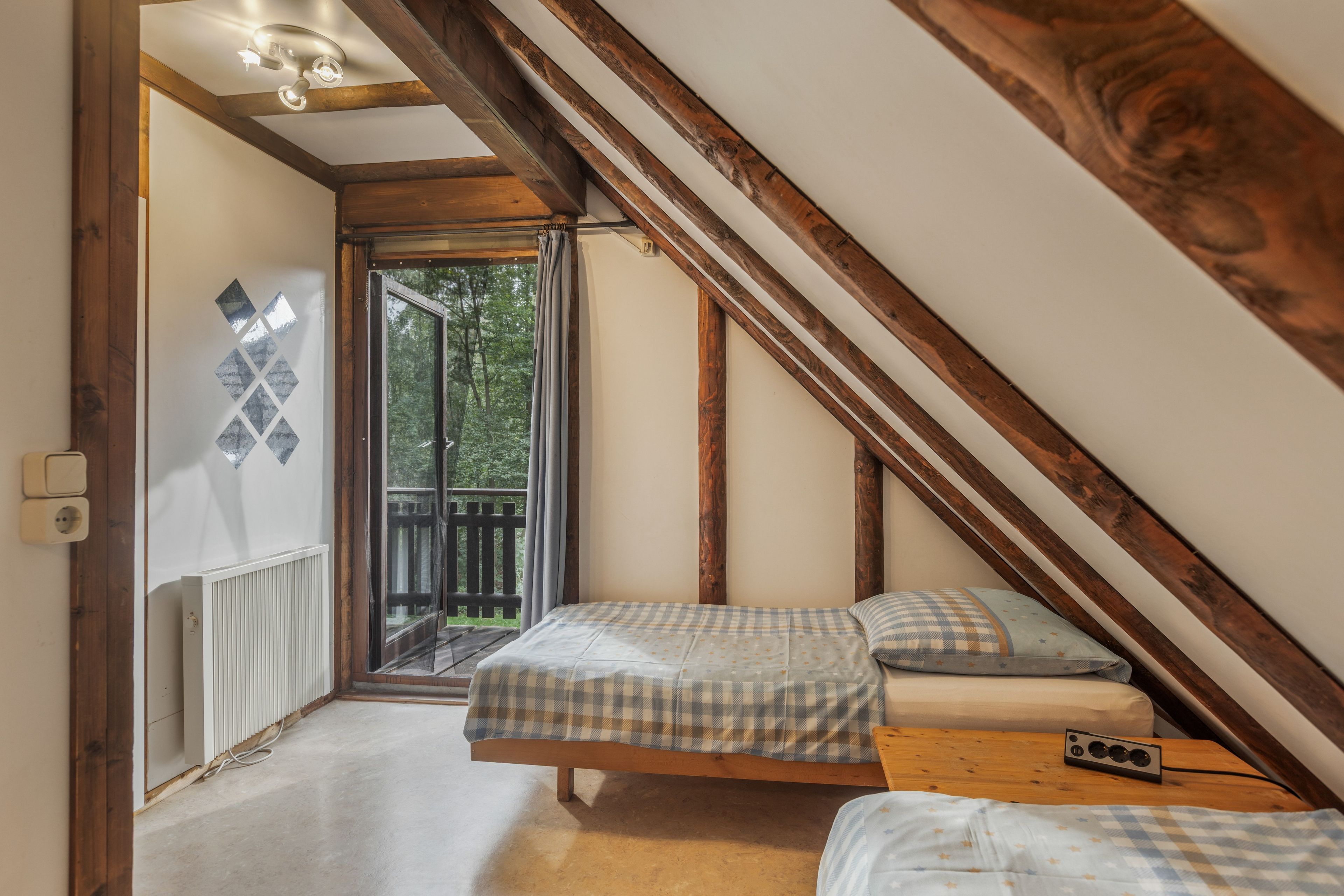 A bedroom with a sloped ceiling, wooden beams, a bed with a checkered bedspread, a bedside table with a radio, and a view of the outdoors through a glass door.