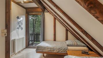 A bedroom with a sloped ceiling, wooden beams, a bed with a checkered bedspread, a bedside table with a radio, and a view of the outdoors through a glass door.