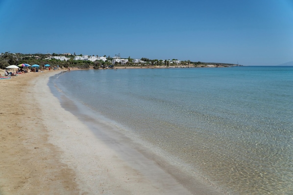A beach with clear water, a sandy shore, and buildings in the background.