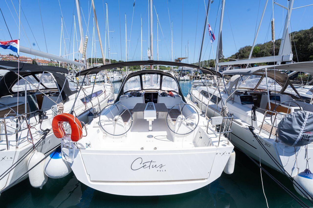 A marina with multiple sailboats docked, including a white boat named 'Cetus Pula' with a life ring and steering wheel visible.