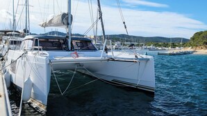 Eine weiße Yacht liegt in einer Marina vor Anker, das Wasser ist ruhig, weitere Boote sind sichtbar und im Hintergrund erstreckt sich eine hügelige Landschaft.