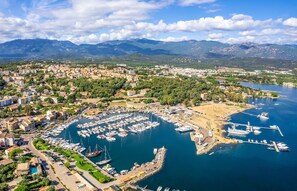 An aerial view of a coastal town with a marina, residential areas, and mountains in the background.