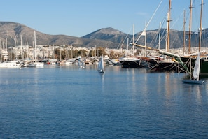 A marina with sailboats, a sailboat with a person on board, and a cityscape with mountains in the background. - Sun-Kissed Sailing Adventure in Islas Sarónicas (Alimos)