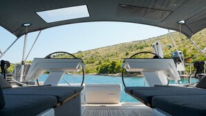 Convés de barco com duas áreas de assento acolchoadas, um leme e vista para um corpo de água e colinas.