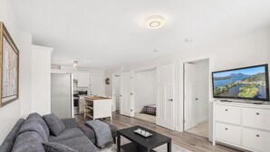 Ein modernes Wohnzimmer mit einer Couch, einem Couchtisch und einem Fernseher. Im Hintergrund ist eine Küchenzeile mit Schränken und einer Mikrowelle zu sehen.