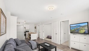A modern living room with a sofa, a coffee table, and a television. There is a kitchen area in the background with cabinets and a microwave. - Modern 2BR Suite Near Lonsdale & Mountains (North Vancouver)