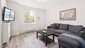 Ein Wohnzimmer mit grauem Sofa, einem schwarzen Couchtisch, einem Fernseher auf einem weißen Schrank und einem gerahmten Bild an der Wand.