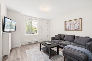 A living room with a grey sofa, a black coffee table, a TV on a white cabinet, and a framed picture on the wall. - Modern 2BR Suite Near Lonsdale & Mountains (North Vancouver)