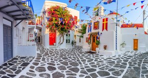A narrow street with white buildings, stone pavement, and colorful flags.