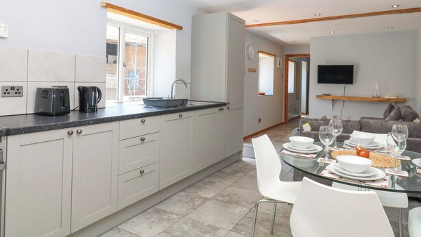 A modern kitchen with a large island, a dining area with a glass table, and a living room with a sofa and TV. - The Milk Shed (Cullompton)