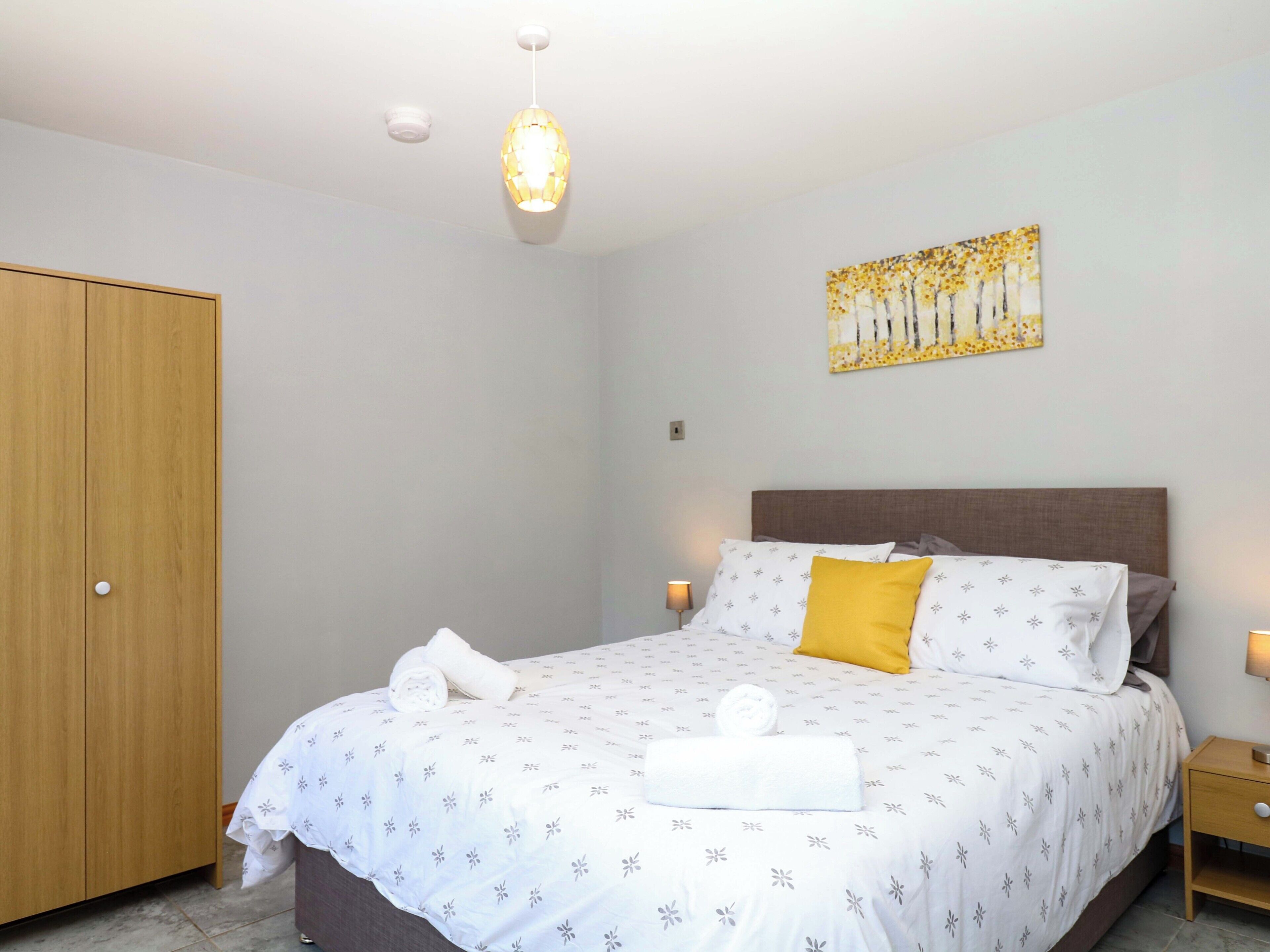 Un dormitorio con cama, mesita de noche, armario de madera, luz de techo y una obra de arte en la pared.