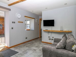 A modern living room with a grey sofa, a mounted TV, a window with a view, and a wooden beam ceiling. - The Milk Shed (Cullompton)