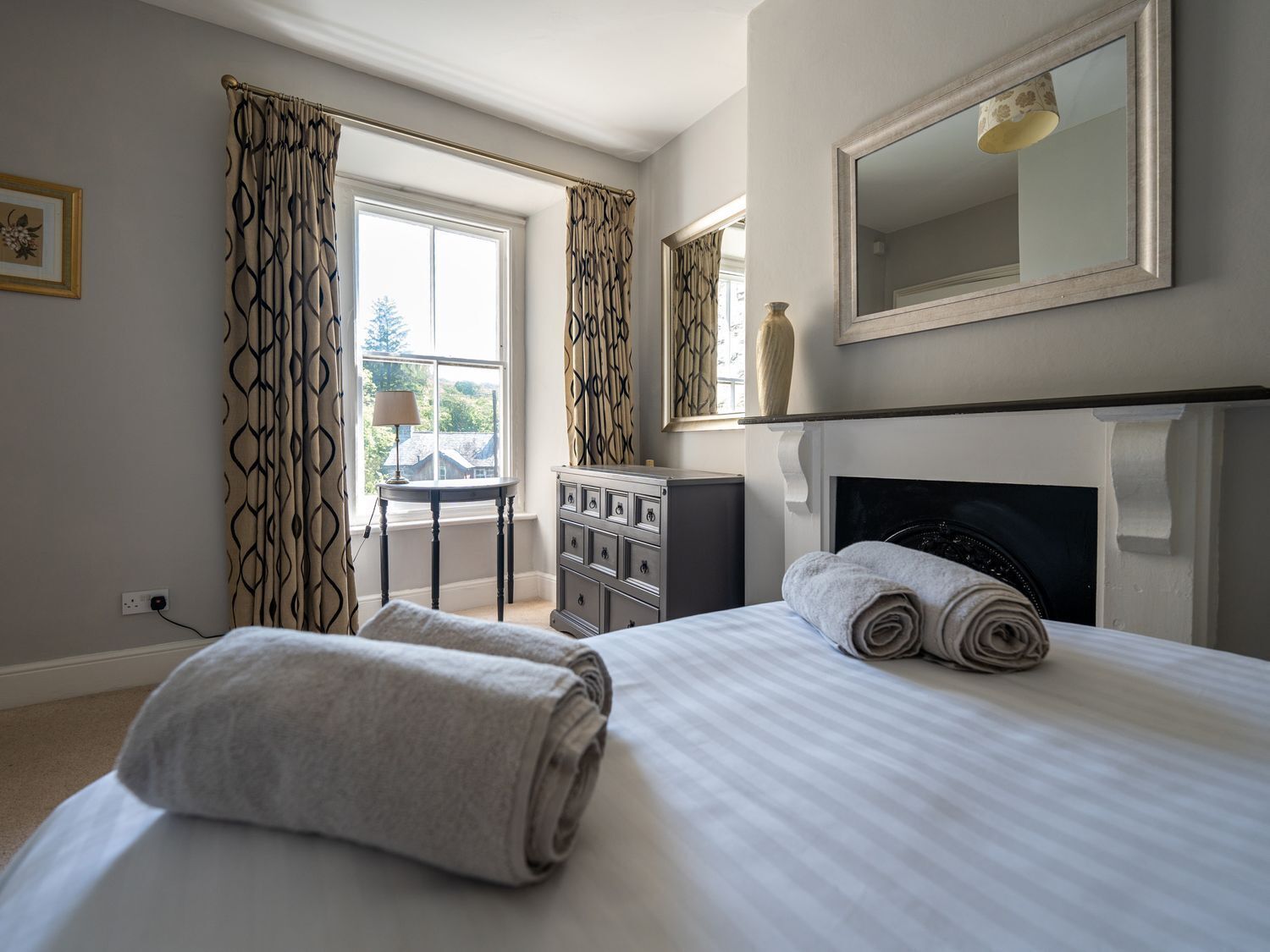 A bedroom with a bed, two rolled towels, a dresser, a mirror, and a fireplace.