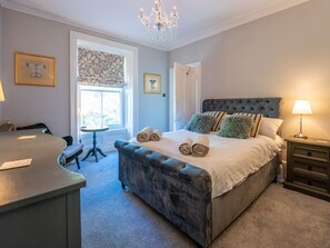 Une chambre à coucher avec une tête de lit capitonnée, un lit agrémenté de serviettes roulées, une lampe de chevet, une fenêtre avec des rideaux à motifs et un lustre.