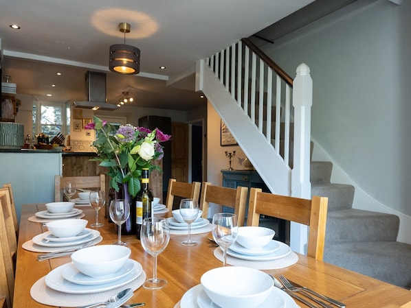 A dining table set for six with white plates, glasses, and cutlery. A vase with flowers is on the table.