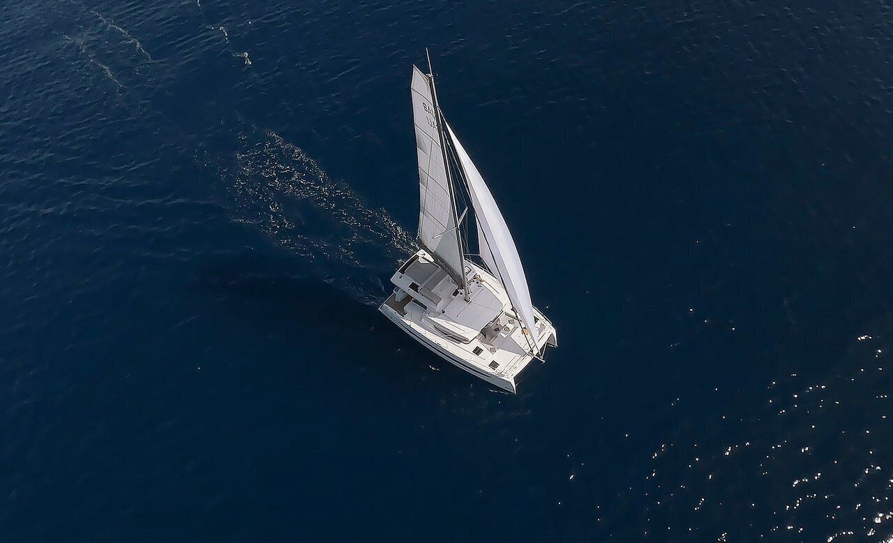 An aerial view of a sailboat with a streamlined hull and a large sail, floating on calm water.