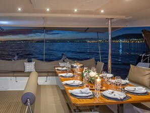 Uma área de refeições em um barco com uma mesa posta para dois, vista para a água e um pôr do sol ao fundo.