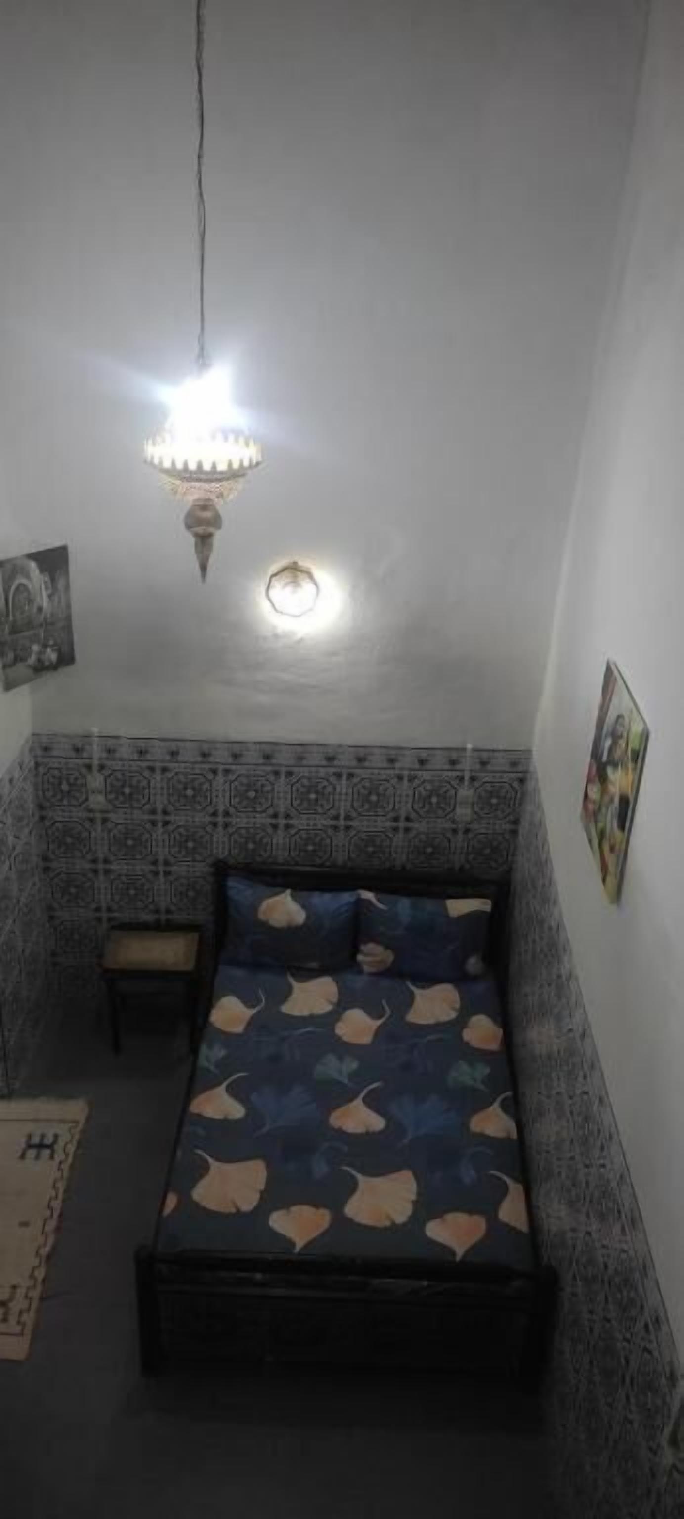 A small room with a bed, a nightstand, a wall-mounted shelf, and a hanging light fixture.