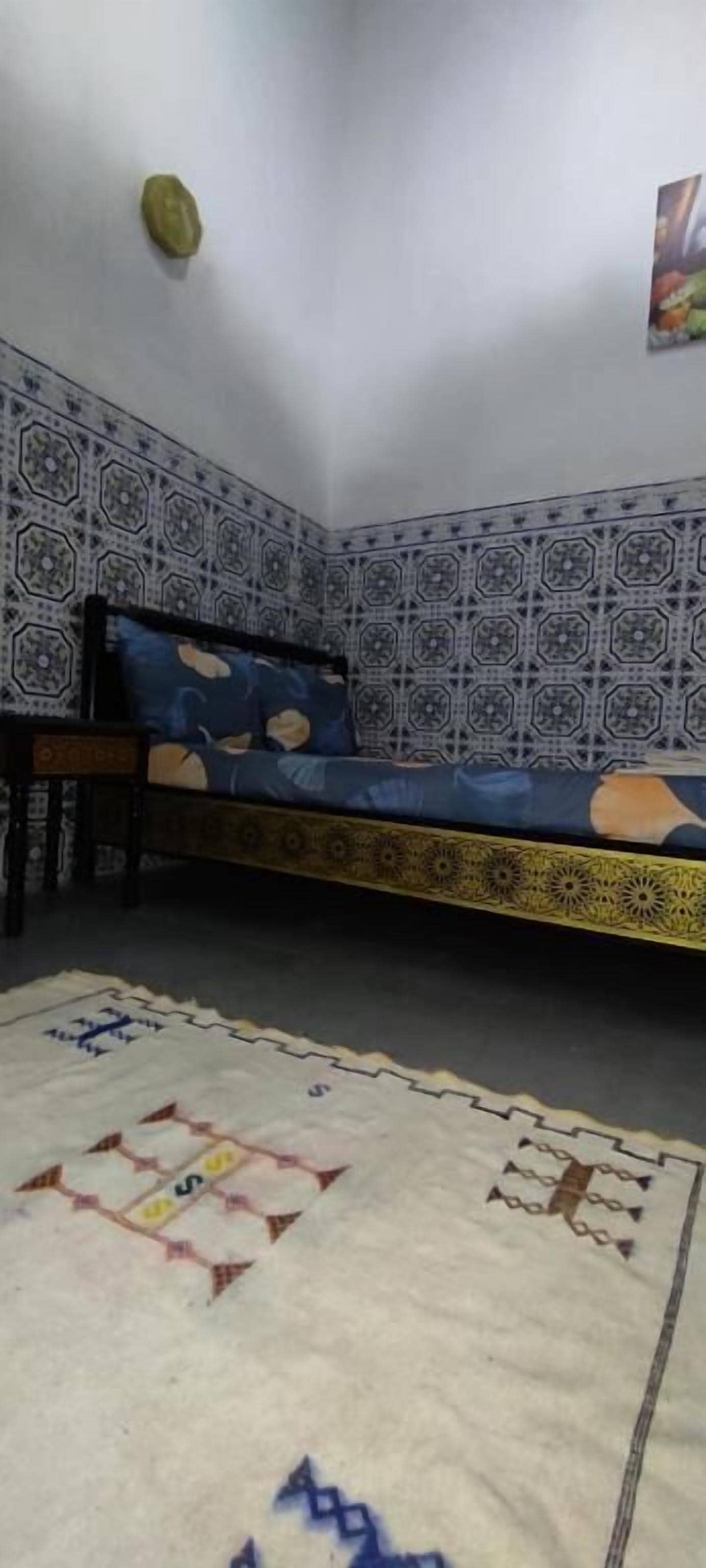 Une pièce comprenant un lit, une petite table, un tapis à motifs et un mur orné de carreaux disposés selon un motif géométrique.