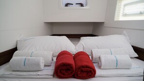 Een net opgemaakt bed met witte kussens, een paar rode handdoeken en een klein raam met uitzicht.
