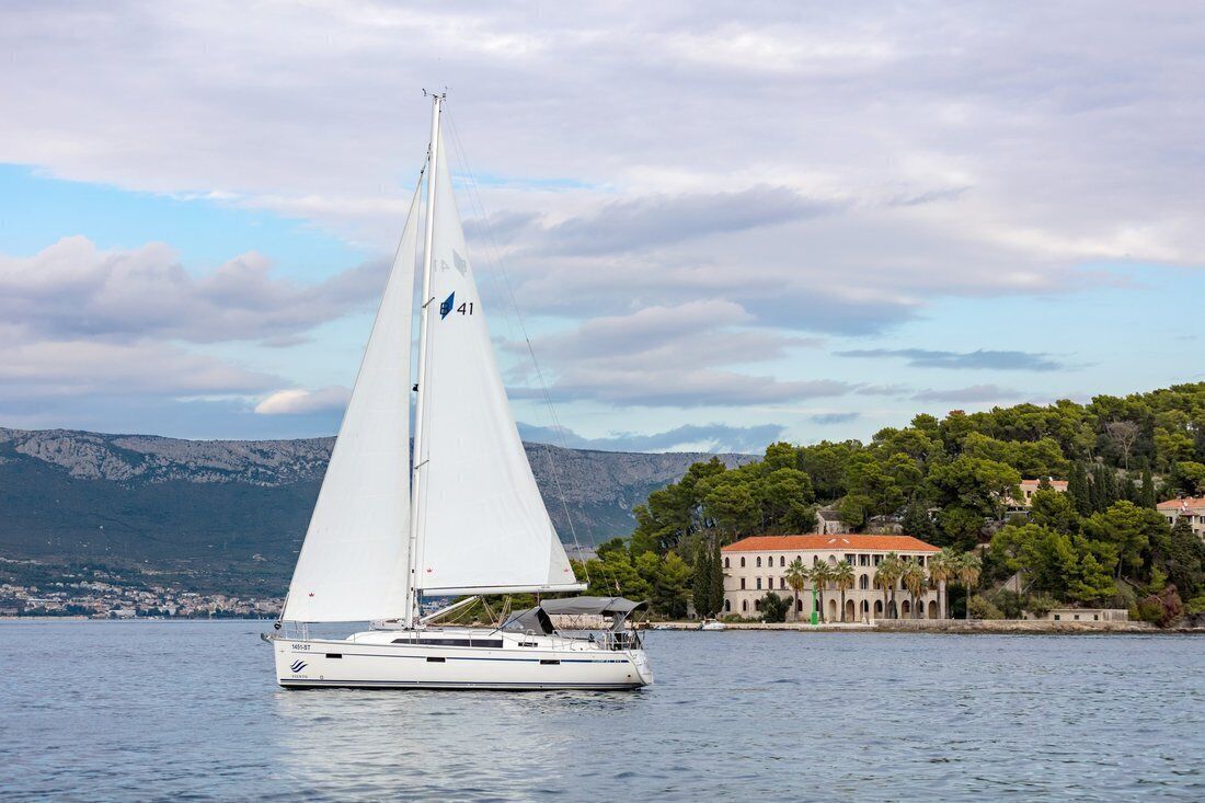 A sailboat on calm waters with a mountainous backdrop and a coastal building.