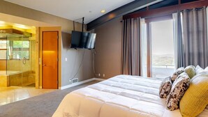 Habitación de hotel con una cama grande, televisor montado en la pared, cabina de ducha de vidrio y vistas al exterior.
