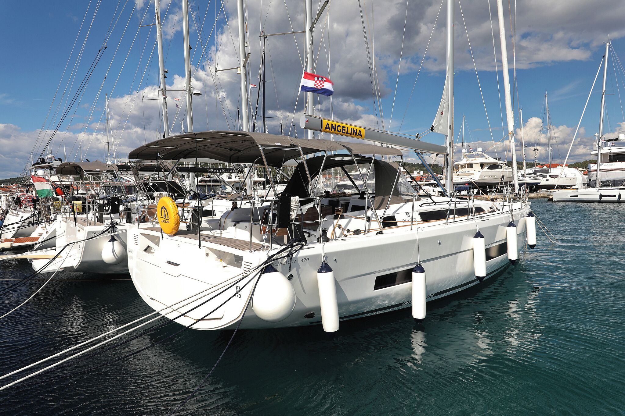 A marina with multiple sailboats docked, including one with a flag and a sign that reads 'ANGELINA.'