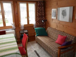 A wooden cabin interior with a bed, a sofa, a chair, a small table, and a window with curtains.
