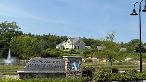 Een stenen bord met de tekst 'Cape Arundel Cottage Preserve' en '1976 Portland Road', een huis op de achtergrond, een fontein en een stenen muur met een muurschildering.