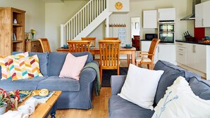 A modern living room with a grey sofa, wooden dining table, and chairs, a staircase, and a kitchen area with white cabinets and a red backsplash. - The Barn at Courtleigh (Kellaton)