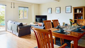 A spacious living area with a dining table set for four, a sofa, a TV, and a bookshelf. - The Barn at Courtleigh (Kellaton)