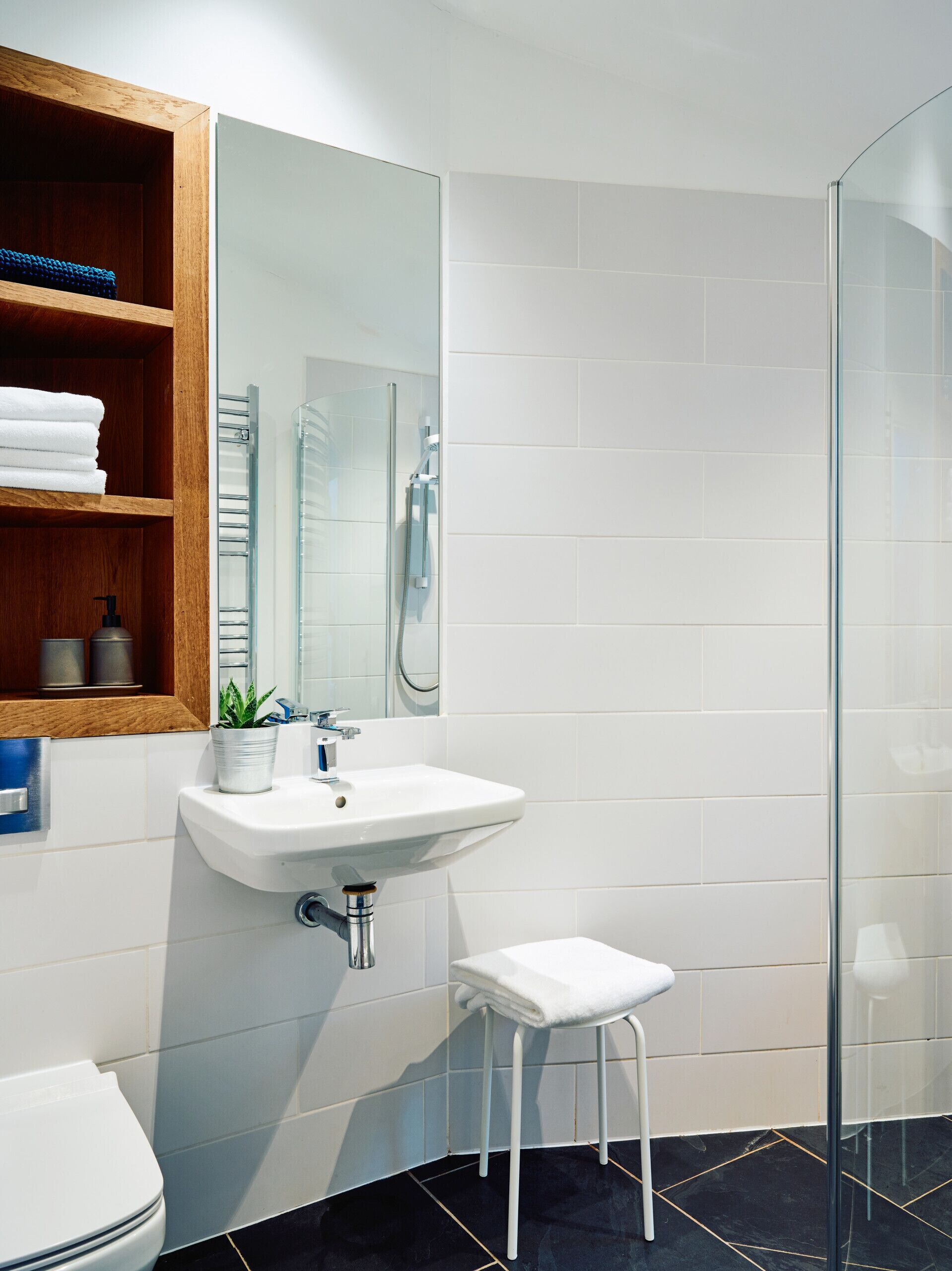 A modern bathroom with a white sink, a glass shower enclosure, a wooden shelf with towels, and a small stool.